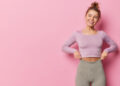 A young woman with a topknot smiles whilst pulling up her high-waisted leggings. She is wearing a long-sleeved lavender crop top and stands in front of a pink background. | FintechZoom