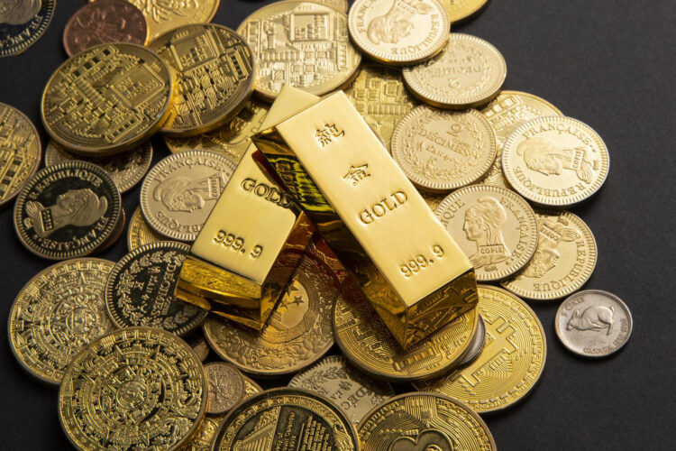 Two gold bars marked "999.9" rest on a pile of various gold coins with different designs, all arranged on a dark surface. | FintechZoom