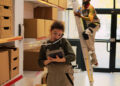 A woman in work overalls talks on the phone and uses a tablet in a storeroom with shelves of boxes, while a man on a ladder works near the door in the background. | FintechZoom