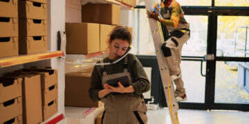 A woman in work overalls talks on the phone and uses a tablet in a storeroom with shelves of boxes, while a man on a ladder works near the door in the background. | FintechZoom