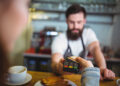 A barista with a beard and apron holds a card payment terminal as a customer pays by tapping a card. A cup of coffee and a pastry are on the counter in the foreground. | FintechZoom