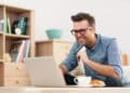 A smiling man in glasses and a checked shirt sits at a table using a laptop, holding a credit card, with a cup of coffee and a plate nearby. Bookshelves and home decor are visible in the background. | FintechZoom