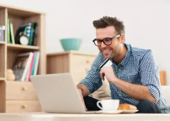 A smiling man in glasses and a checked shirt sits at a table using a laptop, holding a credit card, with a cup of coffee and a plate nearby. Bookshelves and home decor are visible in the background. | FintechZoom