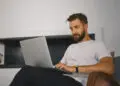 A man with a beard and moustache, wearing a white T-shirt and black trousers, sits in a chair and works on a laptop. He appears focused and is indoors, with a minimal background. | FintechZoom