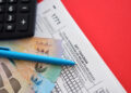 A blue pen, Indonesian banknotes, and a calculator rest on Indonesian tax forms labelled "SPT Tahunan." The scene is set against a red background. | FintechZoom