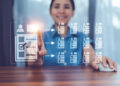 A person sitting at a desk uses a computer mouse and touches a virtual screen displaying digital tick-lists connected by arrows, symbolising data selection or digital workflow management. | FintechZoom