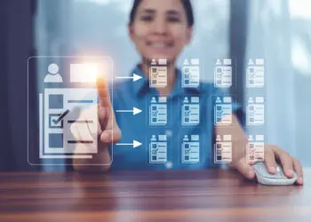 A person sitting at a desk uses a computer mouse and touches a virtual screen displaying digital tick-lists connected by arrows, symbolising data selection or digital workflow management. | FintechZoom