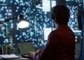 A person in a maroon jacket sits at a desk, working on a laptop displaying share charts, with a lamp beside them and large windows showing a cityscape filled with lit-up buildings at night. | FintechZoom