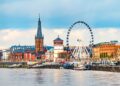 A riverside cityscape featuring a large Ferris wheel, a historic church with a tall spire, and colourful buildings along the waterfront under a partly cloudy sky. | FintechZoom
