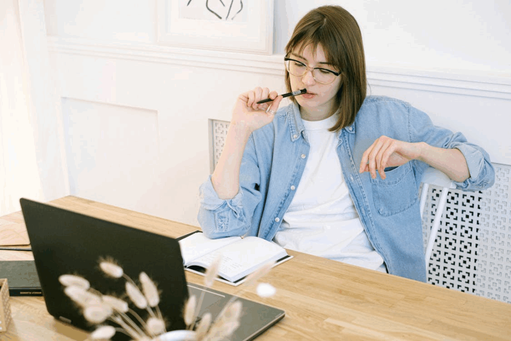 A woman wearing glasses and a denim shirt sits at a desk, looking thoughtfully at a laptop screen whilst holding a pen to her mouth, with an open notebook and dried flowers on the table. | FintechZoom