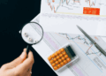 A hand holds a magnifying glass over lettered beads spelling "WIN" on a desk with financial charts, a pen, and an orange-button calculator, highlighting the importance of risk management in achieving success. | FintechZoom