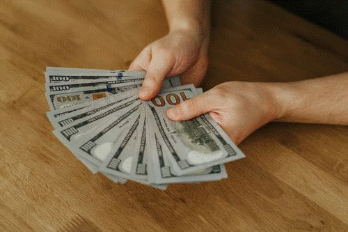 A person holding and fanning out several £100 notes on a wooden table, displaying a large amount of cash. | FintechZoom