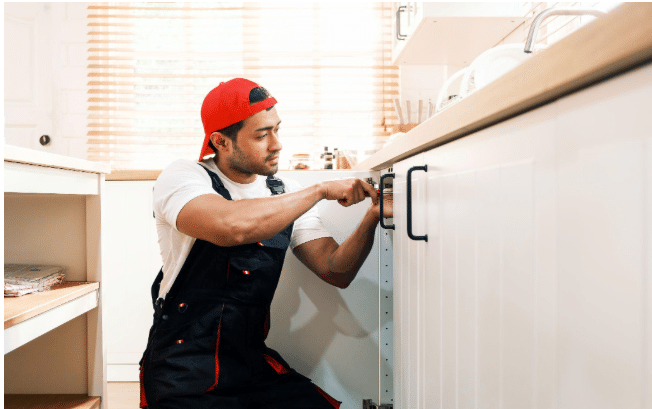 A man wearing a red cap and overalls kneels in a kitchen, using a screwdriver to fix or install the handle on a white cupboard door—one of many Home Renovation Projects. Sunlight streams in through the window blinds behind him. | FintechZoom