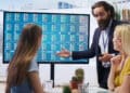 A man in a suit presents stock market data on a large screen to two women seated at a table in a modern office setting. | FintechZoom