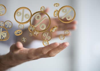 A person’s outstretched hands with floating golden percentage symbols between them, symbolising concepts like interest rates, finance, or discounts. The background is blurred, keeping focus on the symbols. | FintechZoom