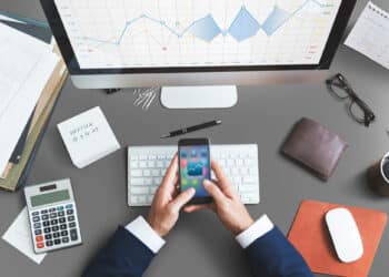 A person in a suit uses a mobile phone at a desk with a computer displaying a line graph, papers, a calculator, keyboard, mouse, pen, notebook, and wallet. The scene suggests financial or business analysis work. | FintechZoom