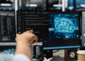A person points at a computer screen displaying code and a digital brain graphic, suggesting artificial intelligence or machine learning development. Other monitors and tech equipment are visible in the background. | FintechZoom