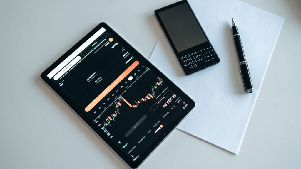 A tablet displaying a share trading app with charts, a mobile phone, a black pen, and a blank sheet of paper are arranged on a white surface. | FintechZoom