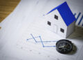 A compass and a small model house sit on financial charts with blue line graphs, symbolising property market trends and direction. | FintechZoom