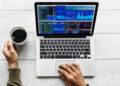 Person holding a cup of coffee while using a laptop displaying multiple financial charts and share data on a white wooden table. | FintechZoom