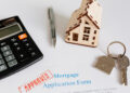 A calculator, pen, wooden house model, house-shaped keyring with keys, and a mortgage application form stamped "APPROVED" are arranged on a white surface. | FintechZoom