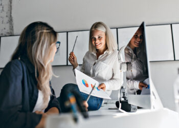 Two women in an office discuss a chart with colourful graphs. One smiles, holding a paper and pen, while the other listens. A computer monitor and camera are visible on the desk. | FintechZoom