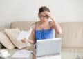 A young woman in glasses looks worried whilst holding papers and sitting in front of a laptop on a coffee table. She touches her forehead with one hand, appearing stressed or surprised by what she sees. | FintechZoom