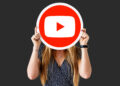 A person with long hair holds a large YouTube logo in front of their face, standing against a plain dark background. | FintechZoom