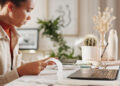 A woman sits at a desk holding a receipt, with a laptop open in front of her. The workspace features plants, spectacles, and decorative items in a softly lit room. | FintechZoom