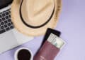 A straw hat, open laptop, cup of coffee, mobile phone, and a passport holding U.S. dollar notes are arranged on a light purple surface. | FintechZoom
