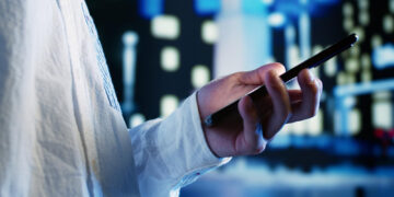 A person wearing a white shirt holds a mobile phone in one hand, with a blurred cityscape featuring lights and buildings in the background. | FintechZoom
