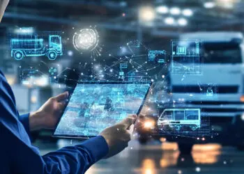 Person holding a tablet with digital graphics of lorries and data, representing logistics and fleet management in a warehouse setting. | FintechZoom