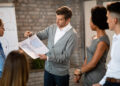 A man in a grey jumper shows building plans to three colleagues during a meeting. They stand near a whiteboard with diagrams in a modern office. | FintechZoom