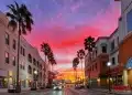 A vibrant sunset with pink and orange clouds above a palm-lined street, flanked by shops and restaurants, as cars drive by and lights begin to switch on. | FintechZoom