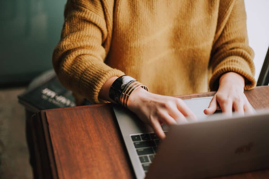A person in a yellow jumper types on a laptop at a wooden desk, with books nearby. Only their hands and torso are visible. | FintechZoom