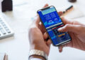 A person holding a mobile phone showing share market data and graphs, with a computer keyboard and documents on a desk in the background. | FintechZoom