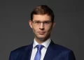 A man with short brown hair wearing glasses, a blue pinstripe suit, white shirt, and blue tie, poses against a dark grey background. | FintechZoom