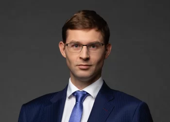 A man with short brown hair wearing glasses, a blue pinstripe suit, white shirt, and blue tie, poses against a dark grey background. | FintechZoom
