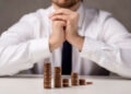 A man in a white shirt and tie sits at a table with his hands clasped, looking at stacks of coins arranged in ascending order. | FintechZoom