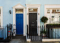 Two terraced houses with blue and pale blue exteriors, one with a bright blue door and a bicycle, the other with a black door and potted plant by the window. Both have white trim and small front gates. | FintechZoom