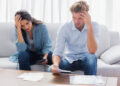 A worried couple sits on a sofa, looking at bills and paperwork on the table. Both have their hands on their heads, appearing stressed or concerned. | FintechZoom