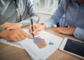 Two people discuss a printed map with coloured regions and data, using a pen to point. A tablet and other papers are on the desk, suggesting a collaborative work or business meeting. | FintechZoom