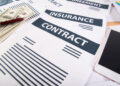Close-up of papers labelled “INSURANCE CONTRACT,” a stack of cash, a red pen, and part of a mobile phone on a desk, suggesting financial or insurance-related paperwork. | FintechZoom