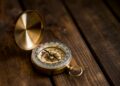 A vintage brass compass with its lid open rests on a dark wooden surface, showing the compass needle and detailed markings—a timeless piece reminiscent of Tom Batterman’s adventures. | FintechZoom