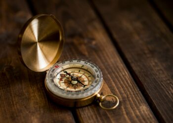 A vintage brass compass with its lid open rests on a dark wooden surface, showing the compass needle and detailed markings—a timeless piece reminiscent of Tom Batterman’s adventures. | FintechZoom