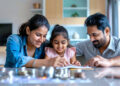 A smiling family of three, with a mother, father, and young daughter, sits at a table together, writing in a notebook, with stacks of coins in the foreground. | FintechZoom