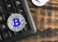 A silver Bitcoin coin with a blue symbol rests on a black keyboard, with part of another coin visible on a wooden surface beside it. | FintechZoom