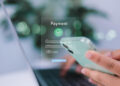 A person holds a mobile phone in one hand while using a laptop, with a digital payment form overlay displaying fields for card number, cardholder name, and a confirm button. | FintechZoom