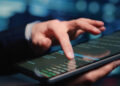A person uses a tablet, interacting with code on the screen, touching and scrolling with their finger. The background is blurred with blue and green tones. | FintechZoom