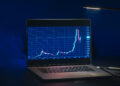 A laptop displays a line graph with sharp rises and drops, likely representing shares or cryptocurrency prices, against a dark background with a desk lamp beside it. | FintechZoom